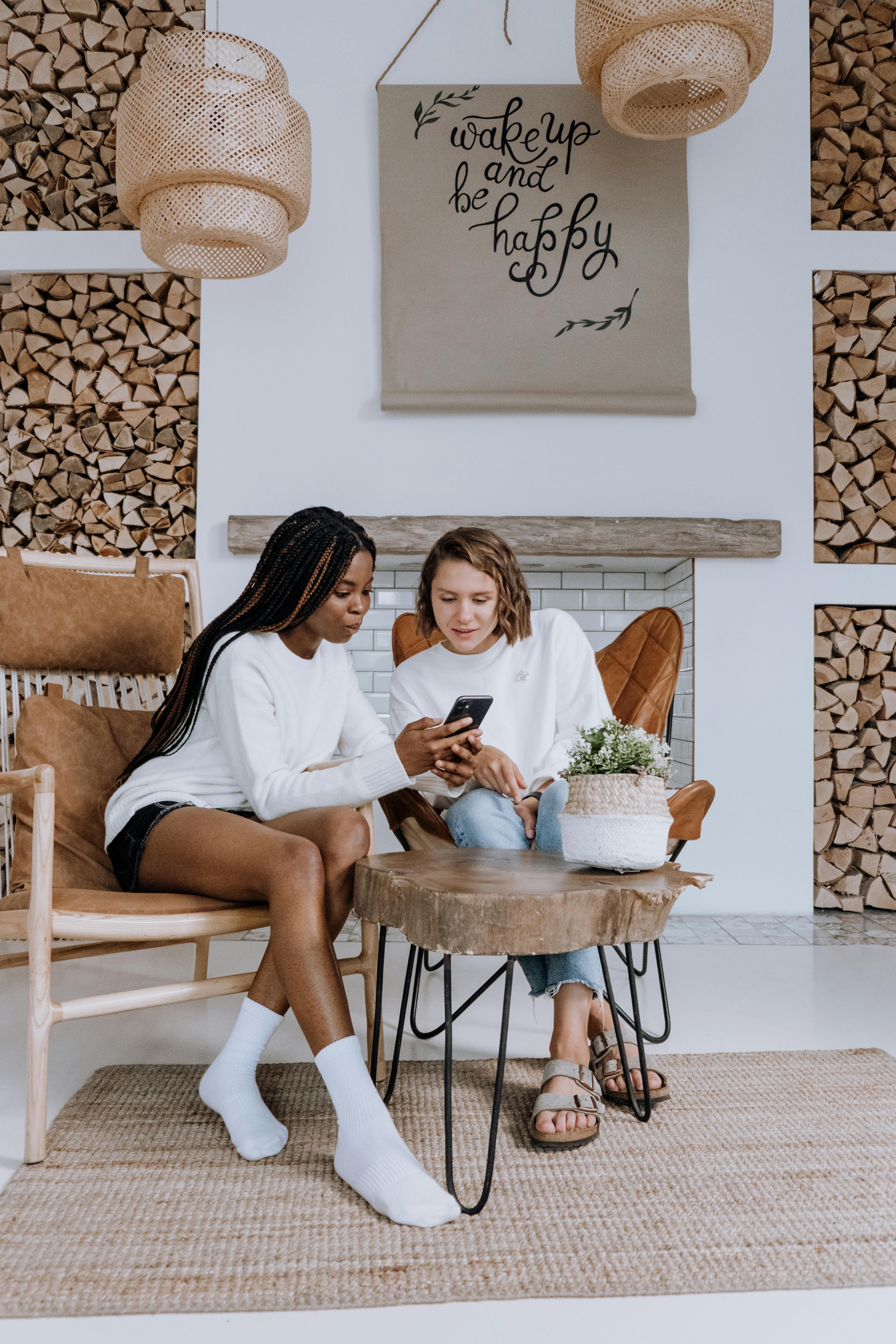 Two women sitting in a cozy room, one showing something on a phone to the other. The decor features wooden accents and a quote on the wall.