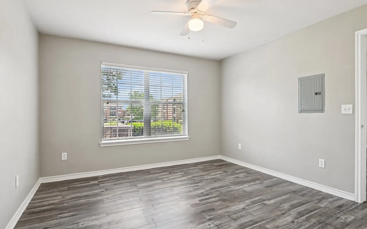 An empty room with light gray walls and brown wooden flooring. A ceiling fan is mounted on the ceiling, and there is a window with blinds letting in natural light. A wall-mounted electrical panel is visible on the right side.