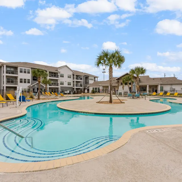 A pool area in an apartment complex featuring a large pool with an island in the center, surrounded by lounge chairs and palm trees under a blue sky with clouds.