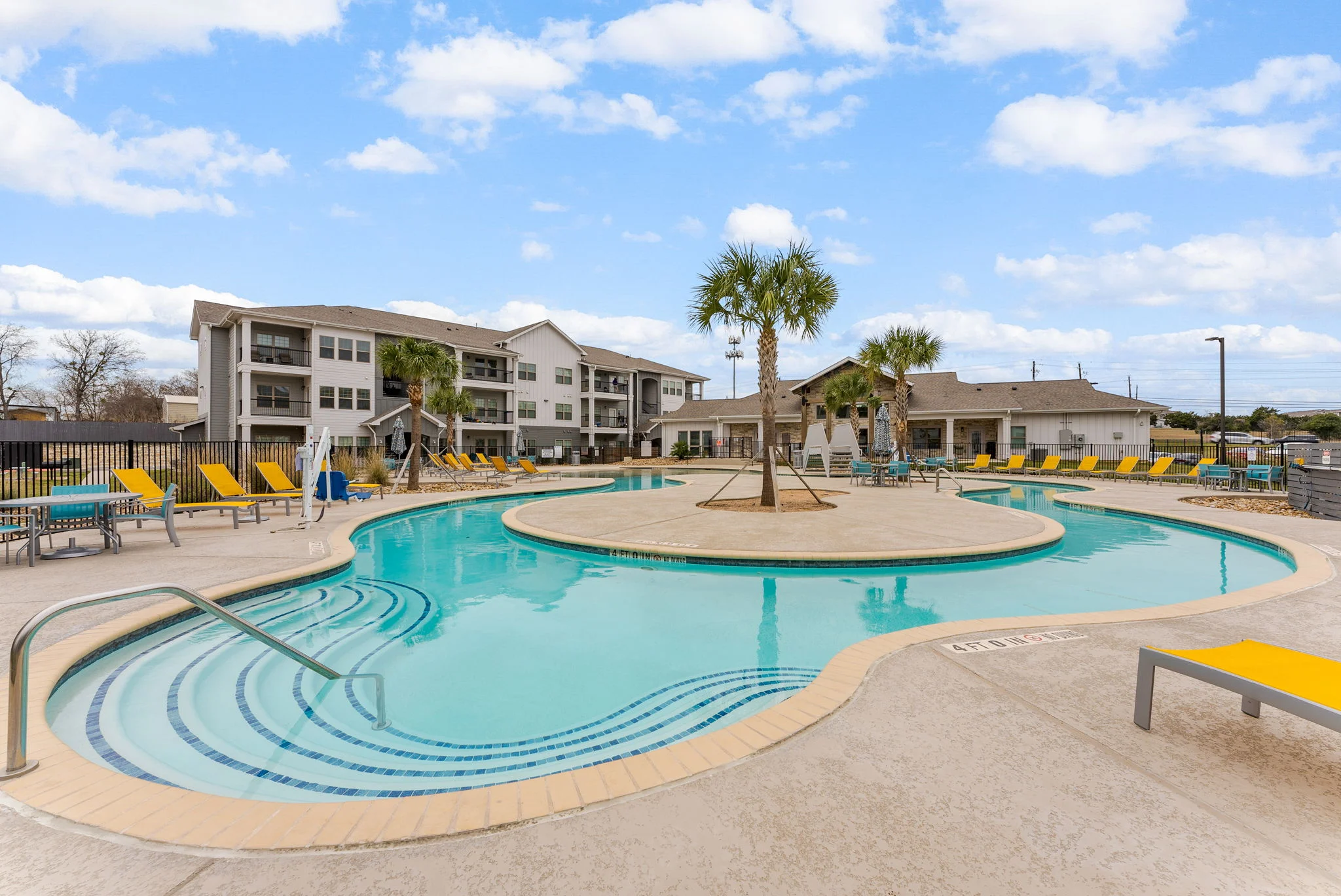 A pool area in an apartment complex featuring a large pool with an island in the center, surrounded by lounge chairs and palm trees under a blue sky with clouds.