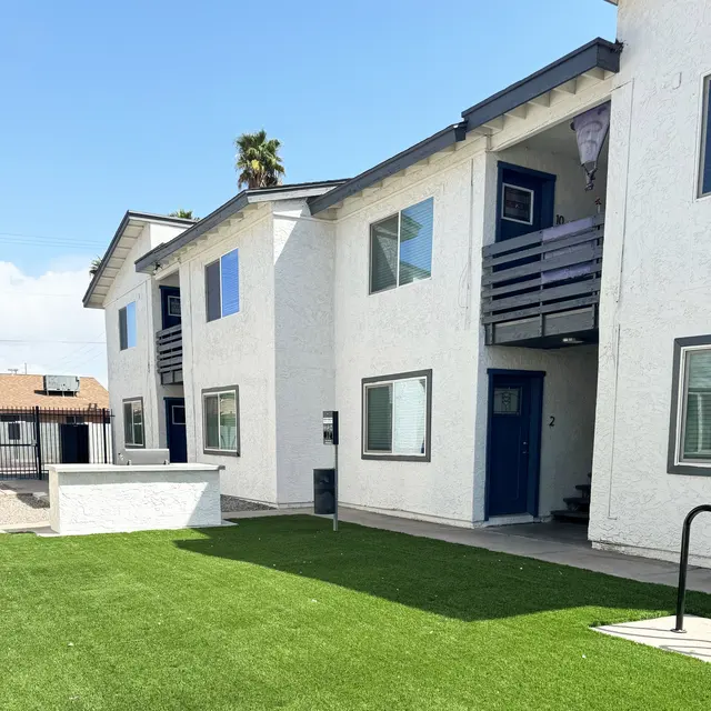 Exterior view of a two-story apartment building with green grass in front. The buildings are white with blue accents, featuring balconies and multiple windows.