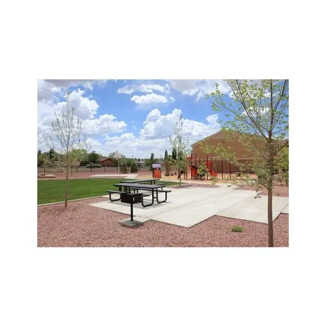 Picnic Area in Park A picnic area in a park with a large table, surrounded by gravel and small trees. In the background, there is a playground structure and a blue sky with clouds.