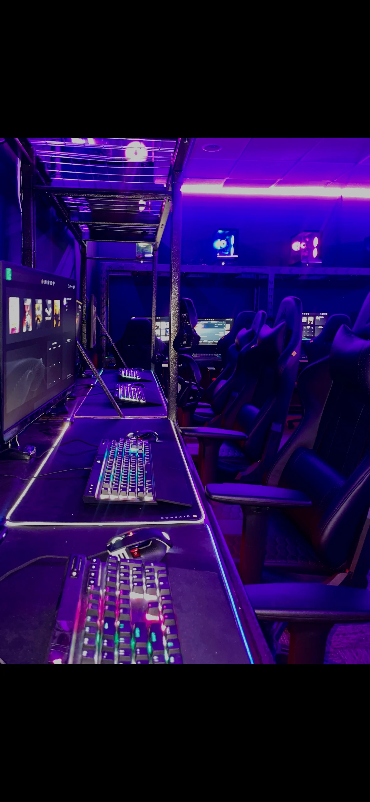 A modern gaming setup with multiple desks and chairs, equipped with high-tech gaming monitors and keyboards. The room is illuminated with purple LED lights, creating a vibrant atmosphere. The chairs are ergonomically designed for comfort.