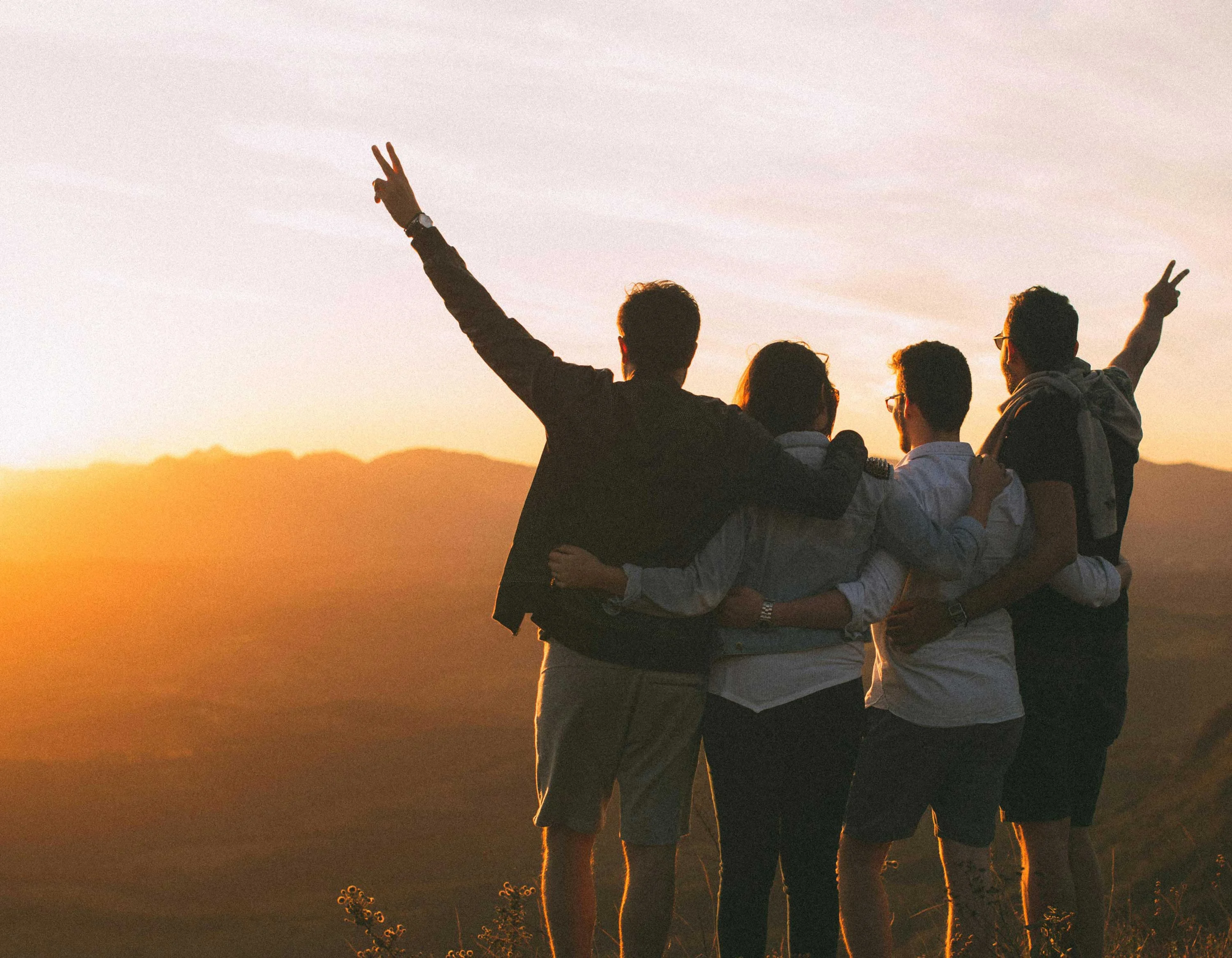 A group of four friends standing together at sunset, with their backs to the camera, enjoying the view and showing peace signs with their hands.
