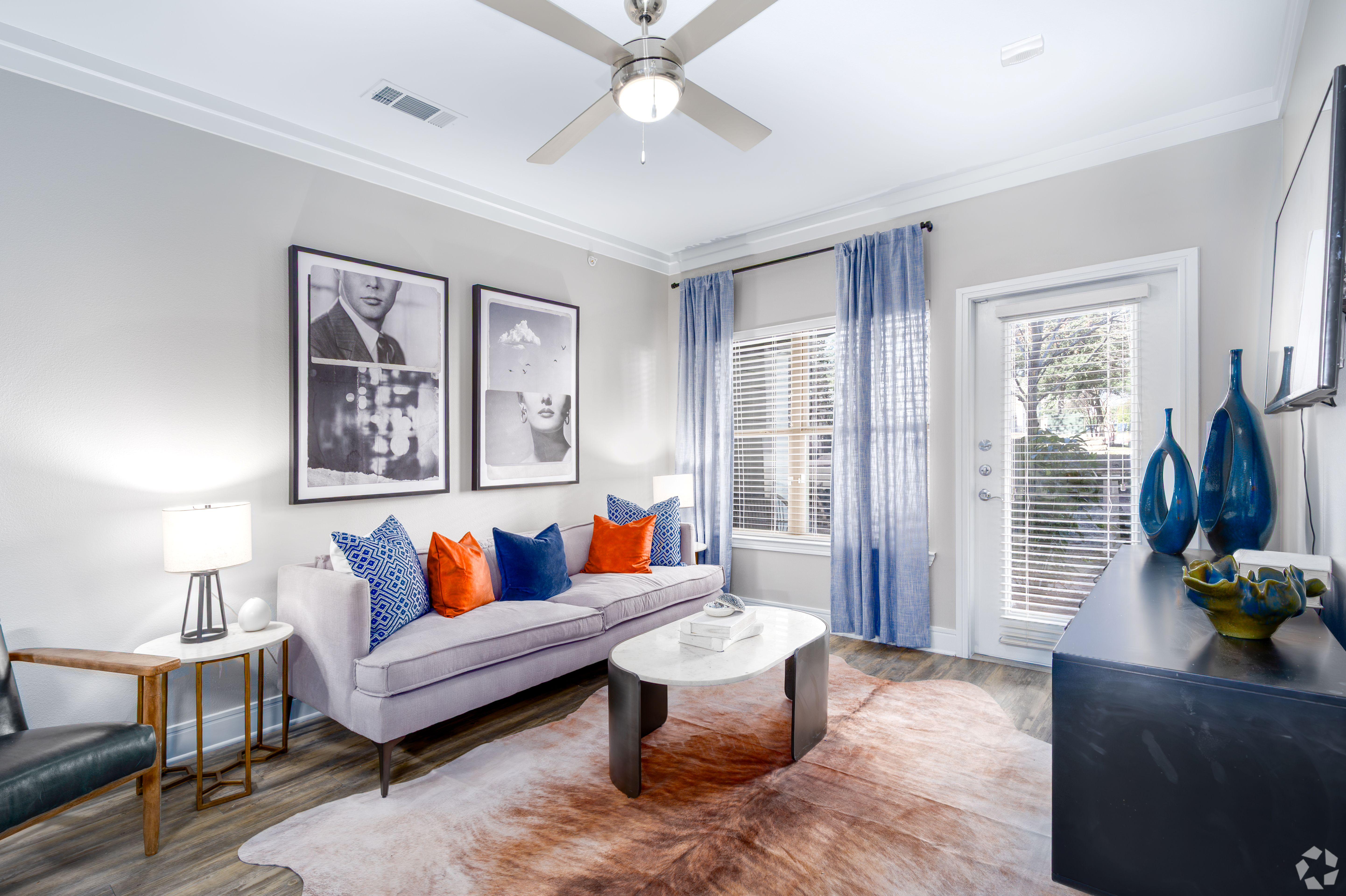 A modern living room featuring a light gray sofa with colorful pillows, a round white coffee table, and a decorative rug on the floor. There are large framed artworks on the wall, and a door leading outside. Sheer blue curtains adorn the window, while a dresser with decorative vases sits against another wall.