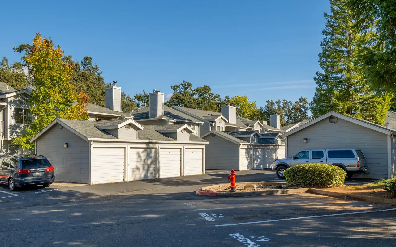 A residential apartment complex featuring several garages, parked vehicles, and trees in the background under a clear blue sky.