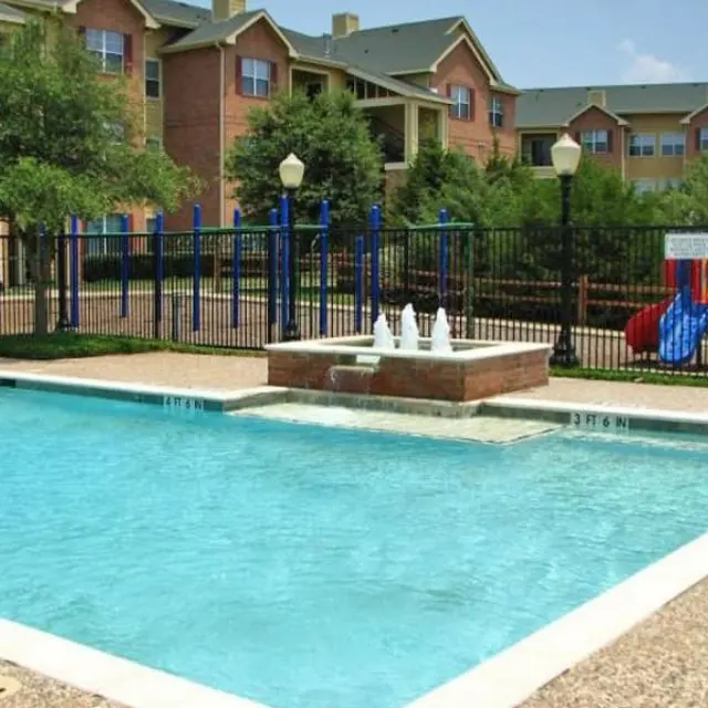 A clear swimming pool with a water fountain in the center, surrounded by a fenced area and greenery. In the background, there are apartment buildings and a children's play area with slides and swings.