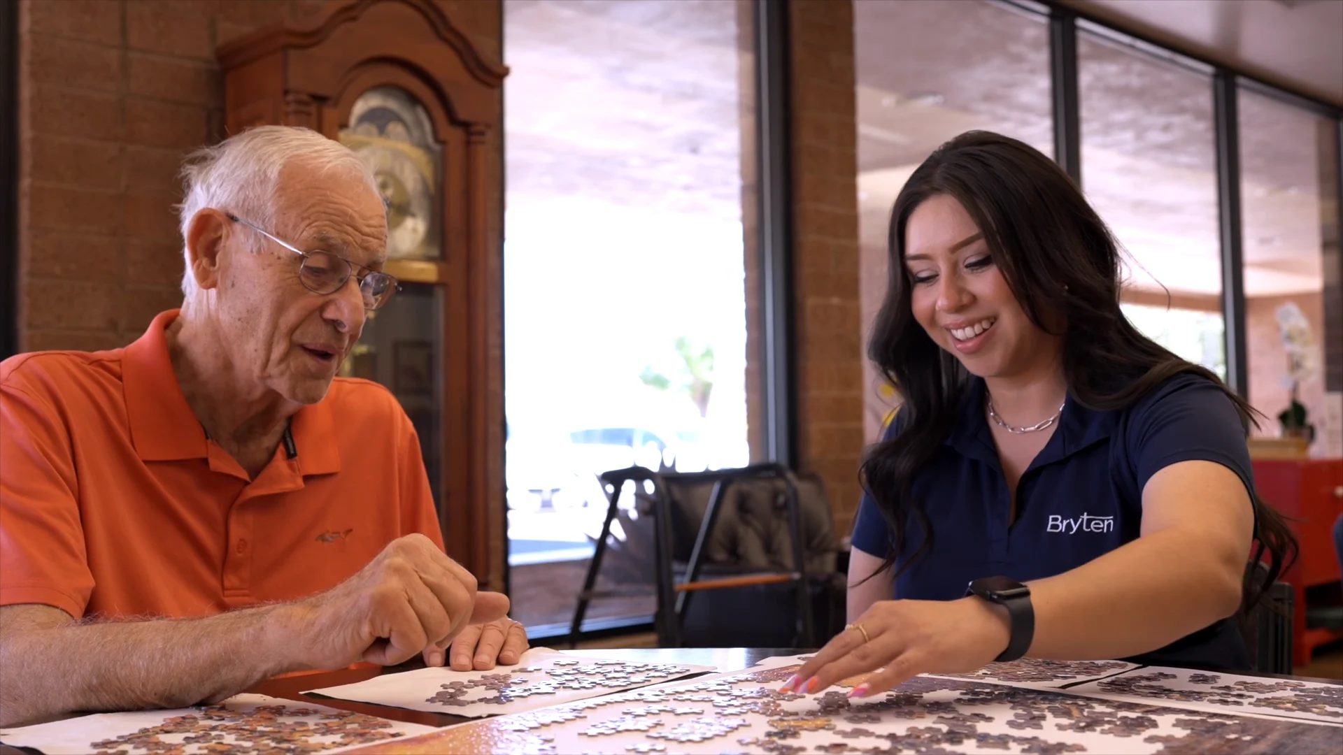An elderly man and a young woman are engaged in a puzzle-making activity at a table in a bright room. The man, wearing an orange shirt and glasses, is focused on the puzzle pieces, while the woman, wearing a navy blue shirt with a logo, is smiling and placing a piece. Natural light pours in through large windows in the background.