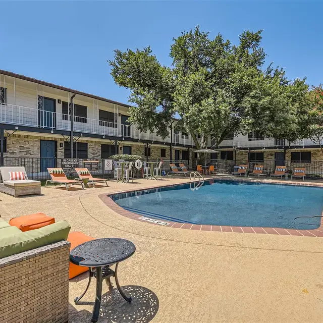 Outdoor pool area surrounded by lounge chairs and an apartment complex. The pool has a few trees around it, and bright orange cushions on the seating area.
