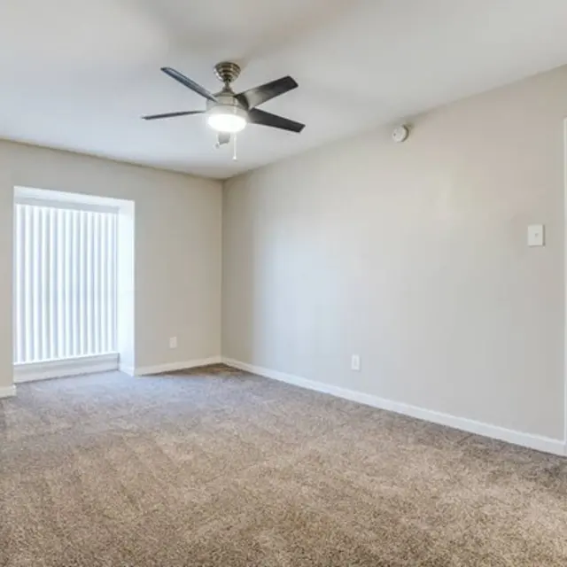A spacious, empty room with light-colored walls and a ceiling fan. There's a window with vertical blinds on one side, and a closed door on the other side. The floor is carpeted in a neutral shade.