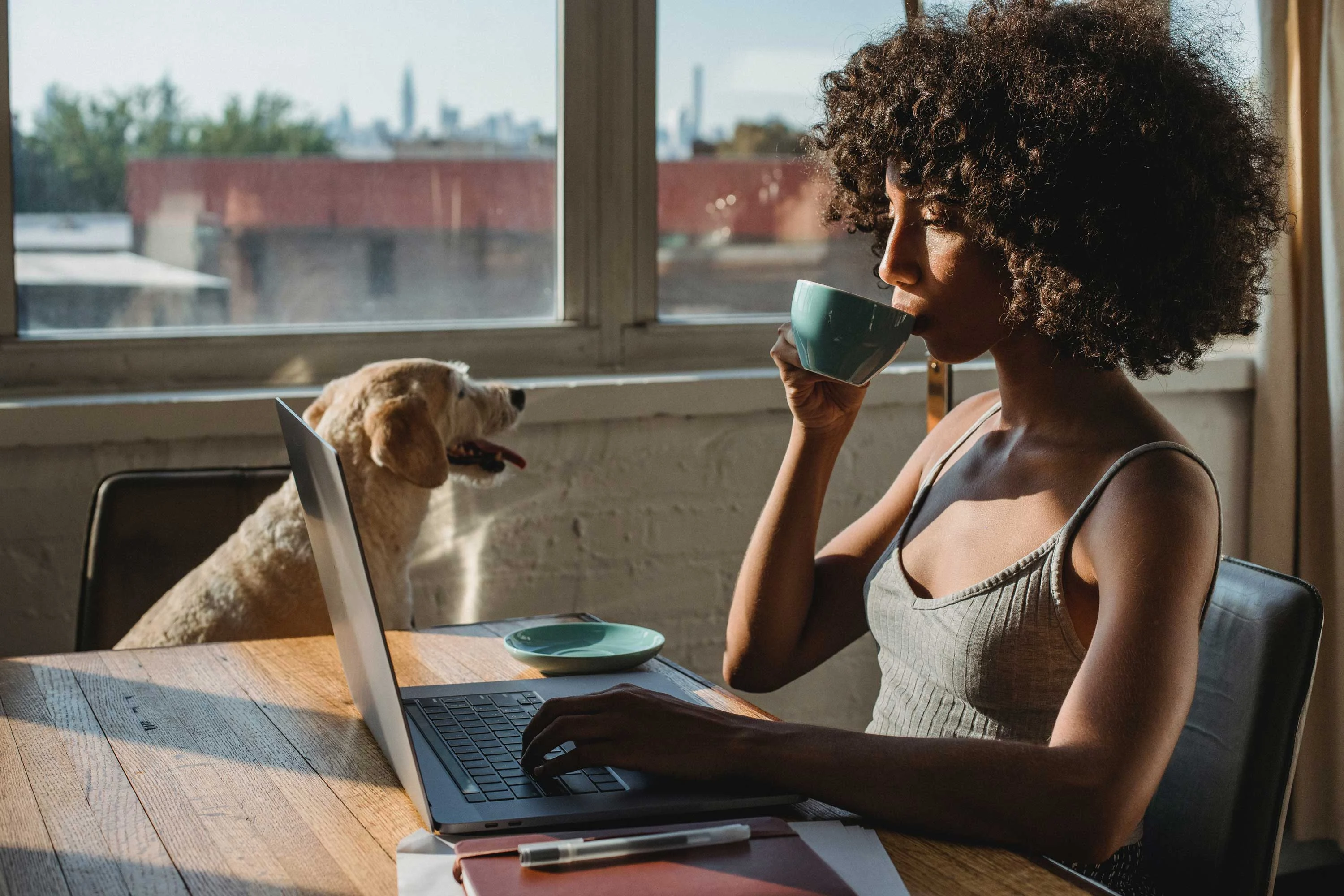 A woman sitting at a table working on a laptop, holding a cup. A dog is sitting beside her near the window.