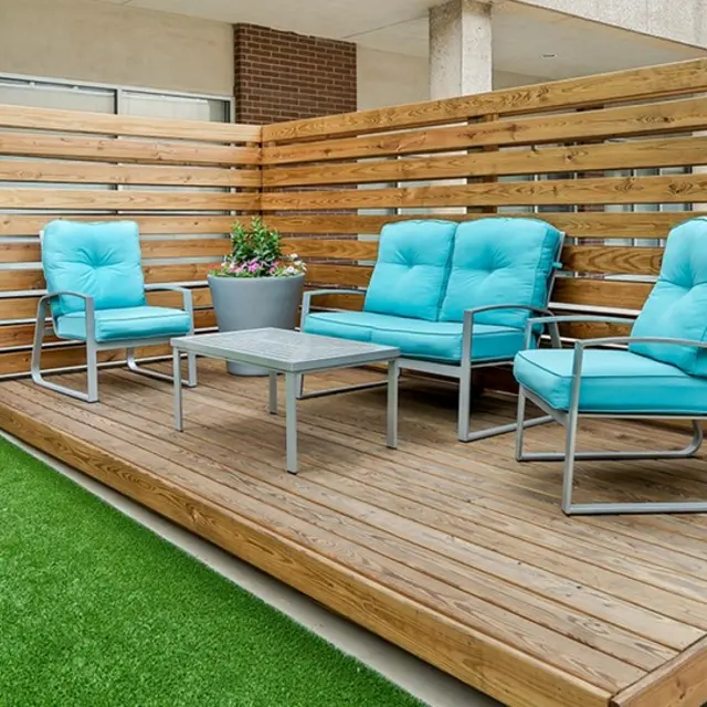 A cozy outdoor seating area featuring two turquoise chairs and a matching loveseat on a wooden deck, with a small table in between and a potted plant on top, surrounded by wooden slats and artificial grass.