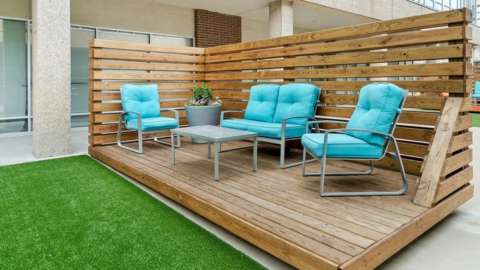 A cozy outdoor seating area featuring two turquoise chairs and a matching loveseat on a wooden deck, with a small table in between and a potted plant on top, surrounded by wooden slats and artificial grass.