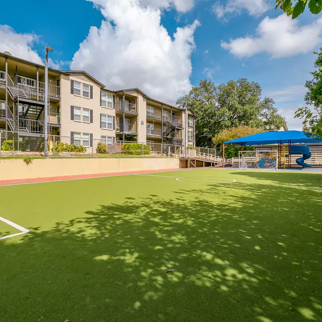 A grassy playground area in front of an apartment complex with a blue slide and a shaded area.