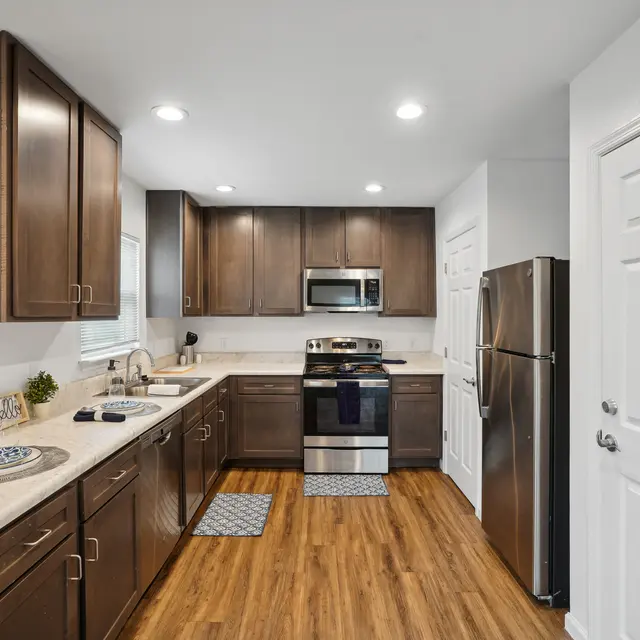 Modern Kitchen Design A modern kitchen featuring dark wood cabinets, stainless steel appliances, and a light countertop. The floor is made of light wooden planks, and the kitchen has a contemporary layout with an open doorway to the right.