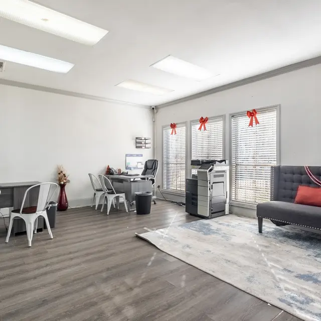 A spacious office interior featuring a seating area with a black couch and decorative pillows, desks with chairs, and a printer. Bright windows with blinds are adorned with red bows. The floor has gray wood-like planks and there’s a rug on the floor.