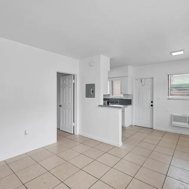 An empty living area of an apartment featuring white walls and tiled flooring, with a small kitchen area visible in the background and a door opening to an exterior.