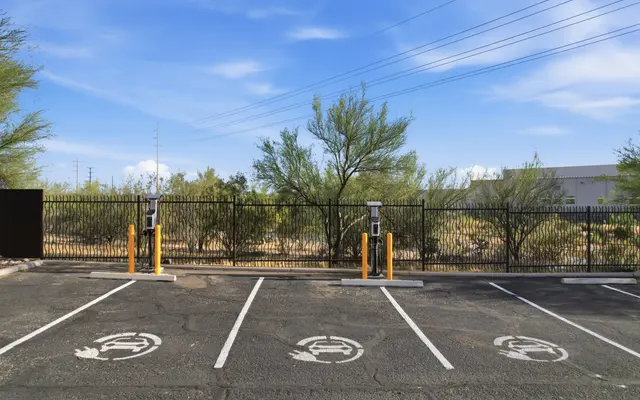 A parking area with two electric charging stations and several empty spaces, bordered by a black metal fence and desert vegetation in the background under a blue sky with clouds.