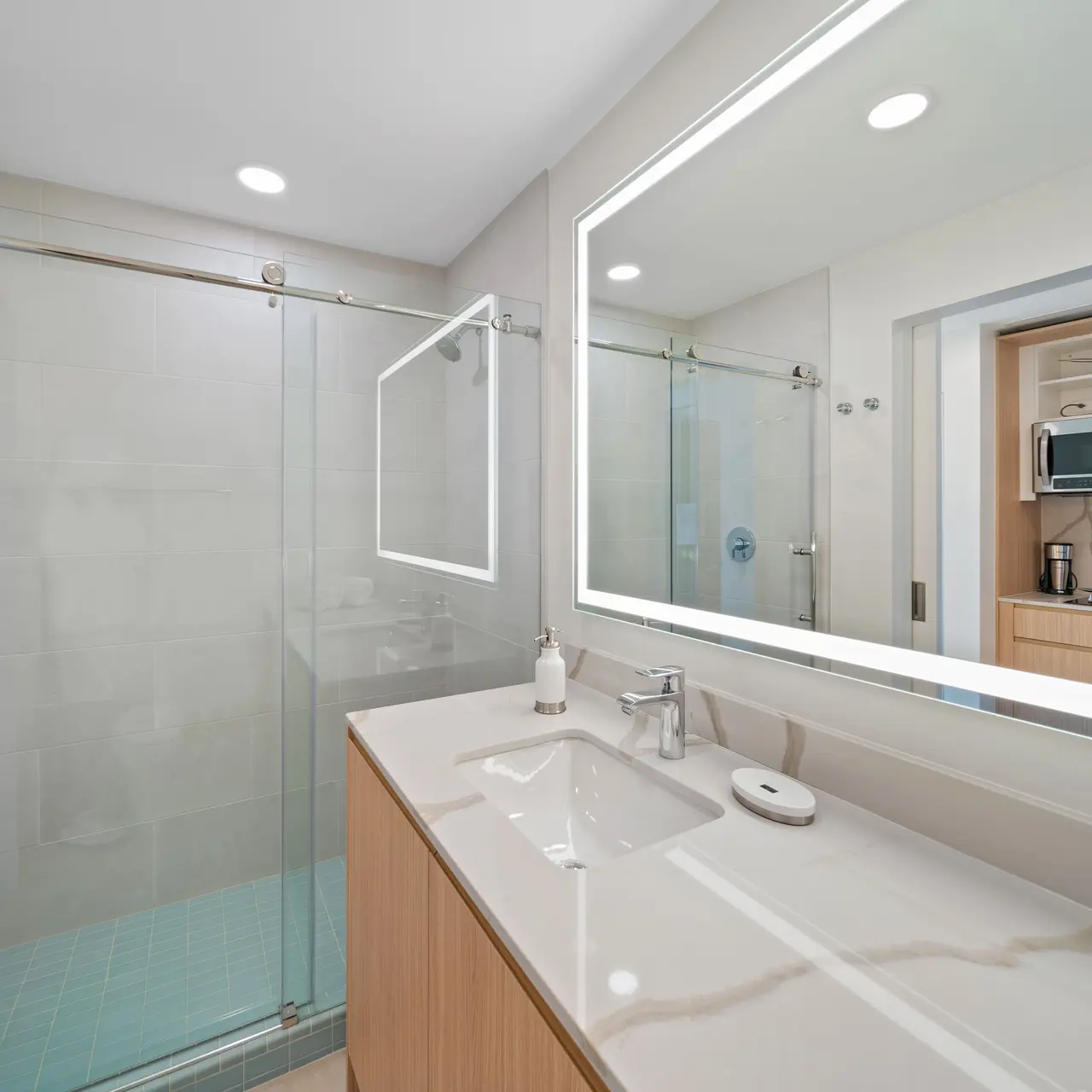 A modern bathroom featuring a walk-in shower with glass doors, a sleek countertop with a sink, and a large illuminated mirror. Soft lighting enhances the clean lines and contemporary design.