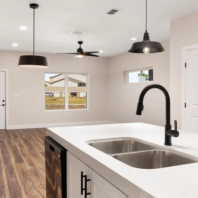 A modern kitchen with a white countertop and stainless steel sink, open to a spacious living area. Pendant lights hang above the counter, and there are large windows on the walls letting in natural light.