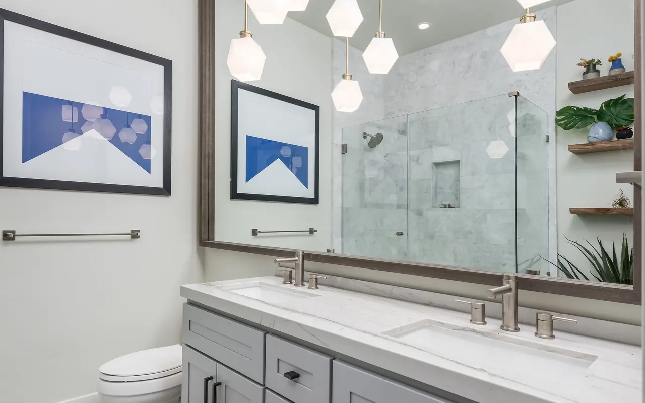 A modern bathroom featuring a double vanity with gray cabinetry, dual sinks, stylish hexagonal pendant lights, and a large mirror reflecting the interior. Two framed artworks are mounted on the wall next to a toilet, with a glass shower in the background and decorative plants on shelves.