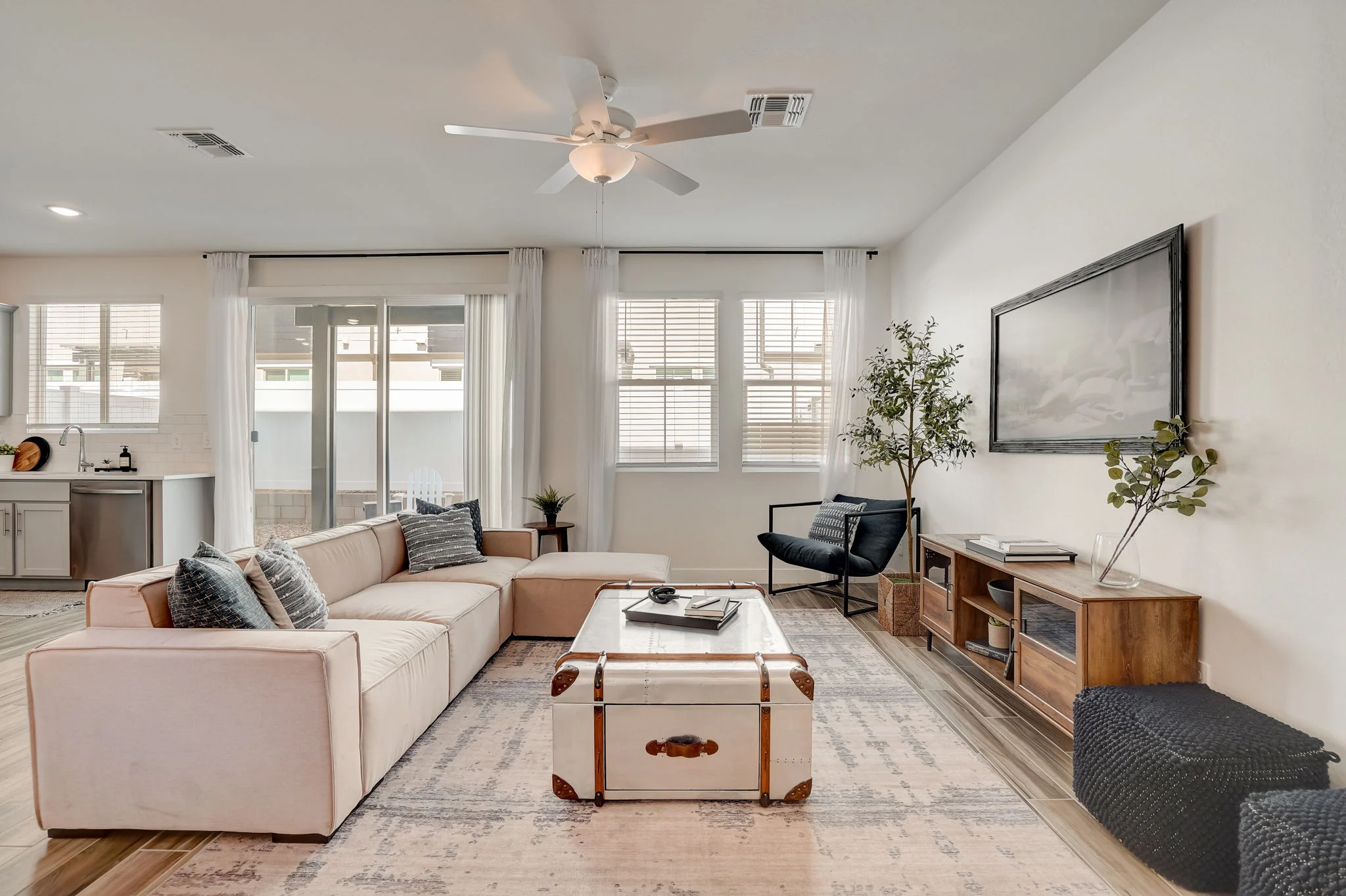 A modern living room featuring a beige sectional sofa, a wooden coffee table, and large windows with sheer curtains. There is a black chair, a small plant, and a flat-screen TV on a wooden console.