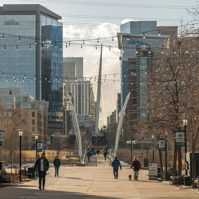 A walkway in downtown Denver, lined with trees and modern skyscrapers, featuring a bridge in the background and people walking.