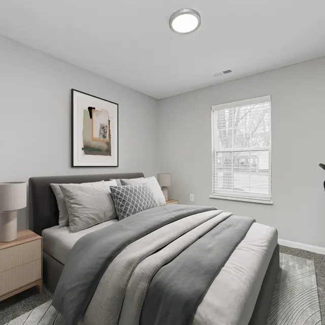 A modern bedroom featuring a neatly made bed with gray bedding, a bedside table with a lamp, a window allowing natural light, and a decorative plant in the corner.