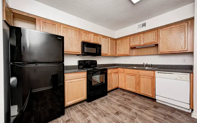 A view of a modern kitchen featuring wooden cabinets, black appliances, and a dishwasher. The flooring is wood-like, and the walls are painted white.