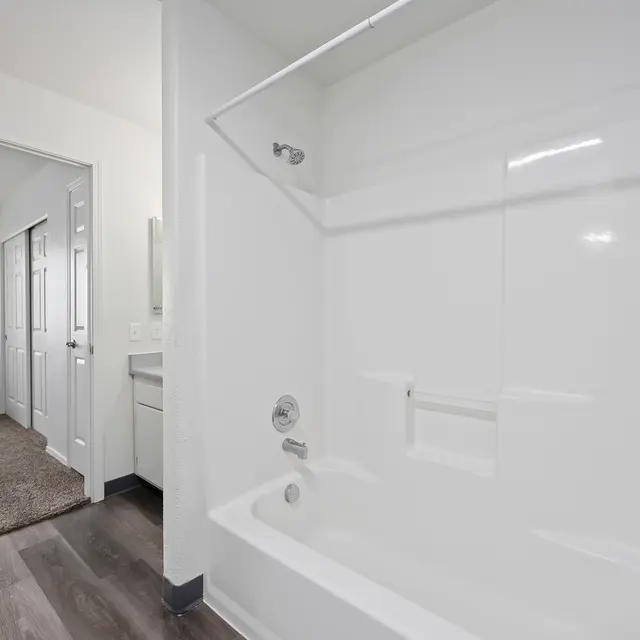 Modern Bathroom Interior A clean and bright bathroom featuring a standalone bathtub with a shower. The walls are painted white, and the floor is made of dark laminate wood. In the background, there is a view into an adjacent room with beige carpeting and doorway.