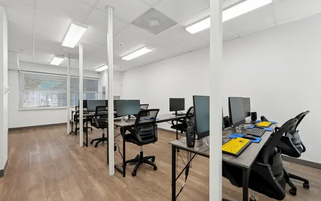 A modern office space featuring several desks with computers and ergonomic chairs, illuminated by bright overhead lights, with a large window providing natural light.