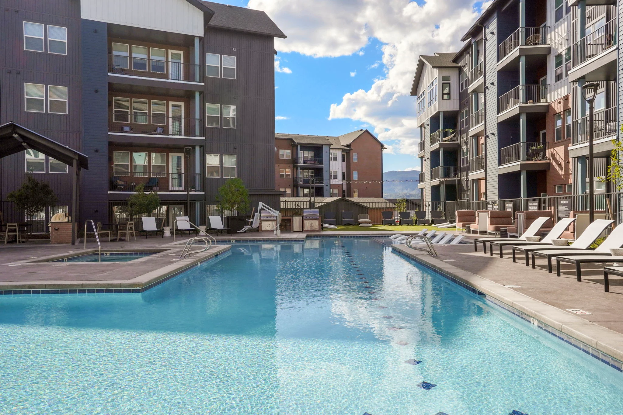 A luxury apartment complex featuring a swimming pool surrounded by lounge chairs and modern buildings in the background with a blue sky and clouds overhead.