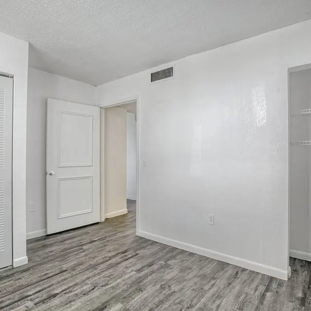 An empty room featuring light-colored walls, a gray wooden floor, a closed closet with sliding doors, and a doorway leading to another room. A small shelf is visible on the right wall.