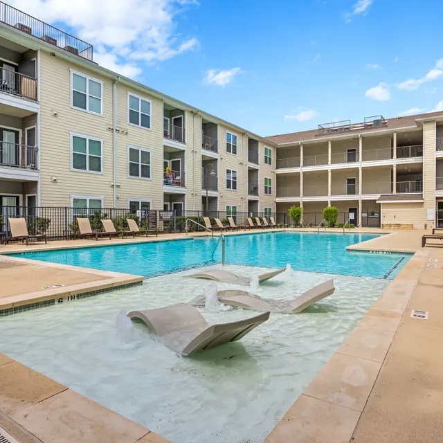 A view of a residential apartment complex featuring a swimming pool. The pool is surrounded by lounge chairs and overlooks several apartment balconies. The sky is clear with a few clouds.