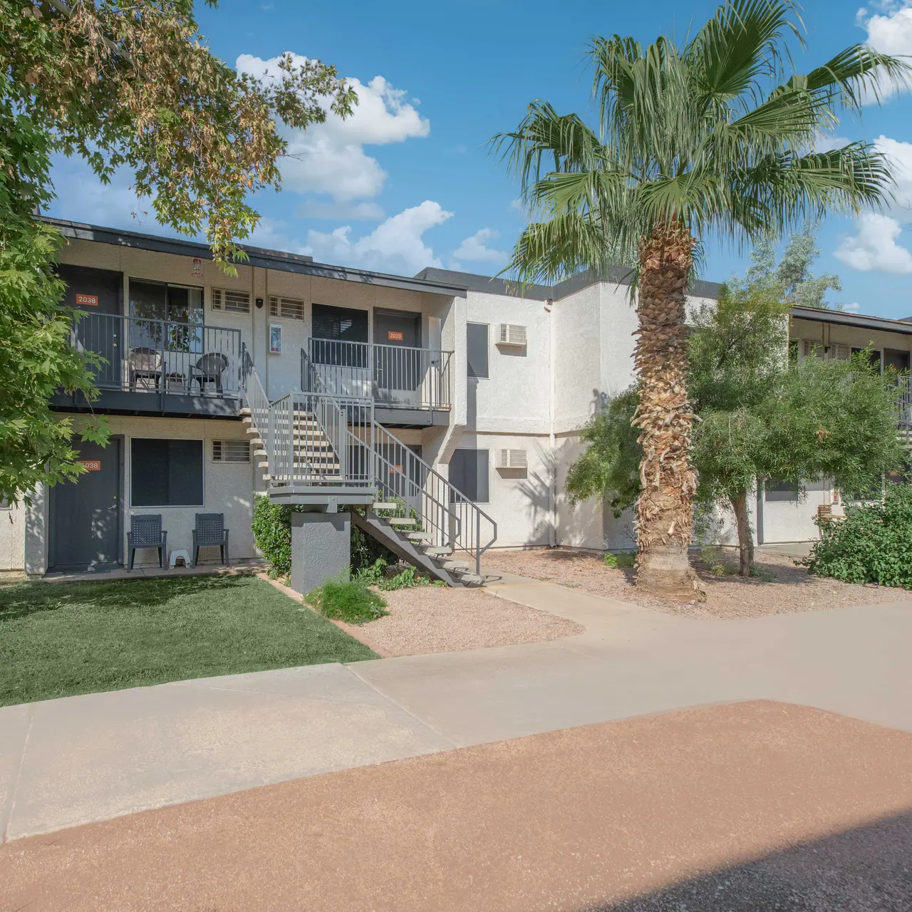 A view of a two-story apartment complex with balconies, surrounded by palm trees and greenery under a clear blue sky.