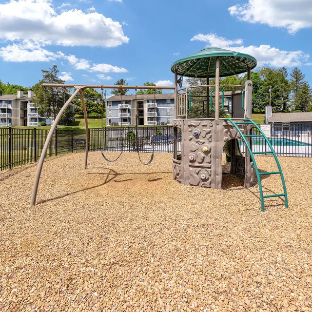 A playground featuring a climbing structure and swings, surrounded by a grassy area and buildings in the background on a sunny day.