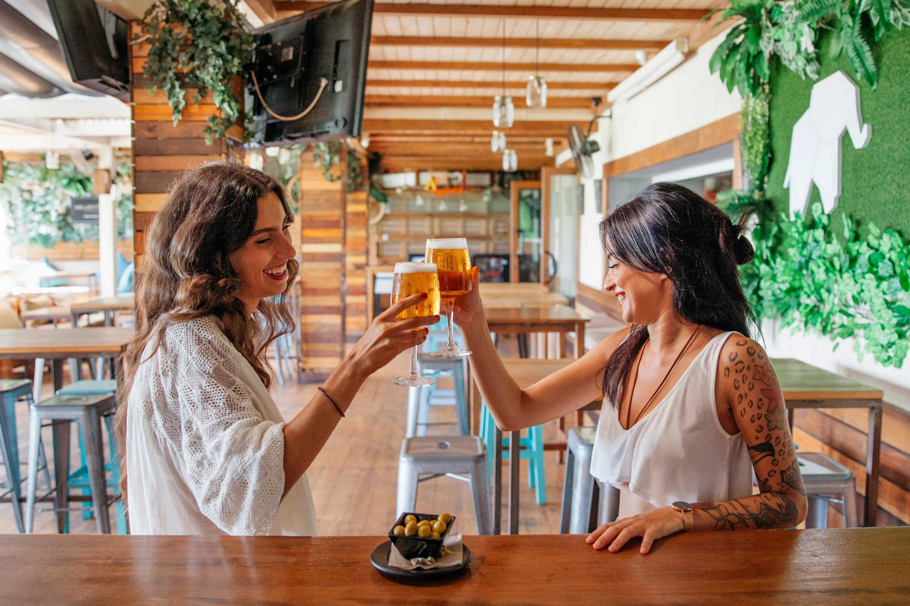 Two women are cheering with drinks in a stylish bar setting, surrounded by greenery and wooden decor.
