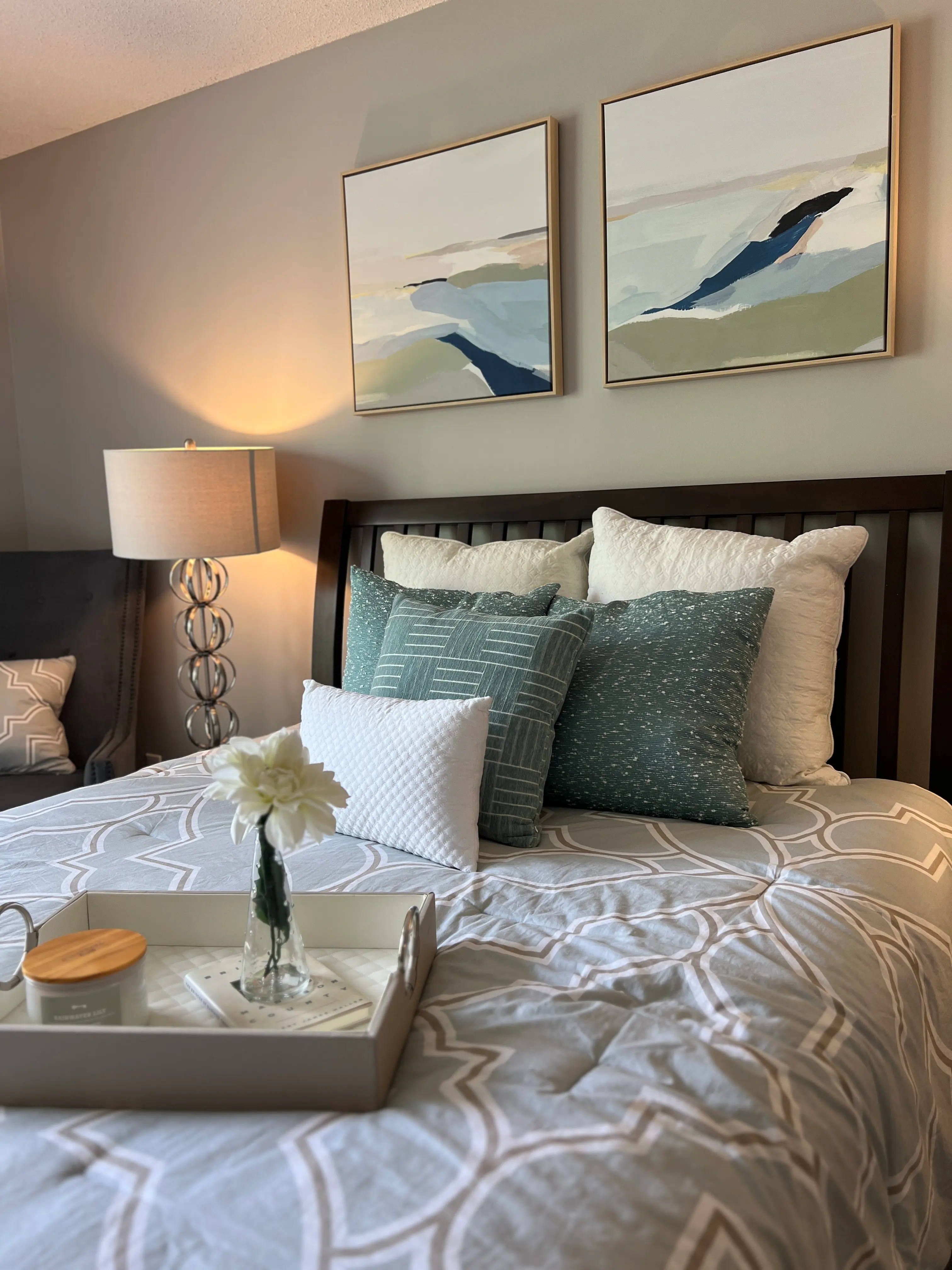 Modern Cozy Bedroom Decor A cozy bedroom featuring a wooden bed with patterned bedding, decorative pillows, and a tray with a flower and snacks on the bed. Two abstract paintings hang on the wall above the bed, illuminated by a nearby lamp.