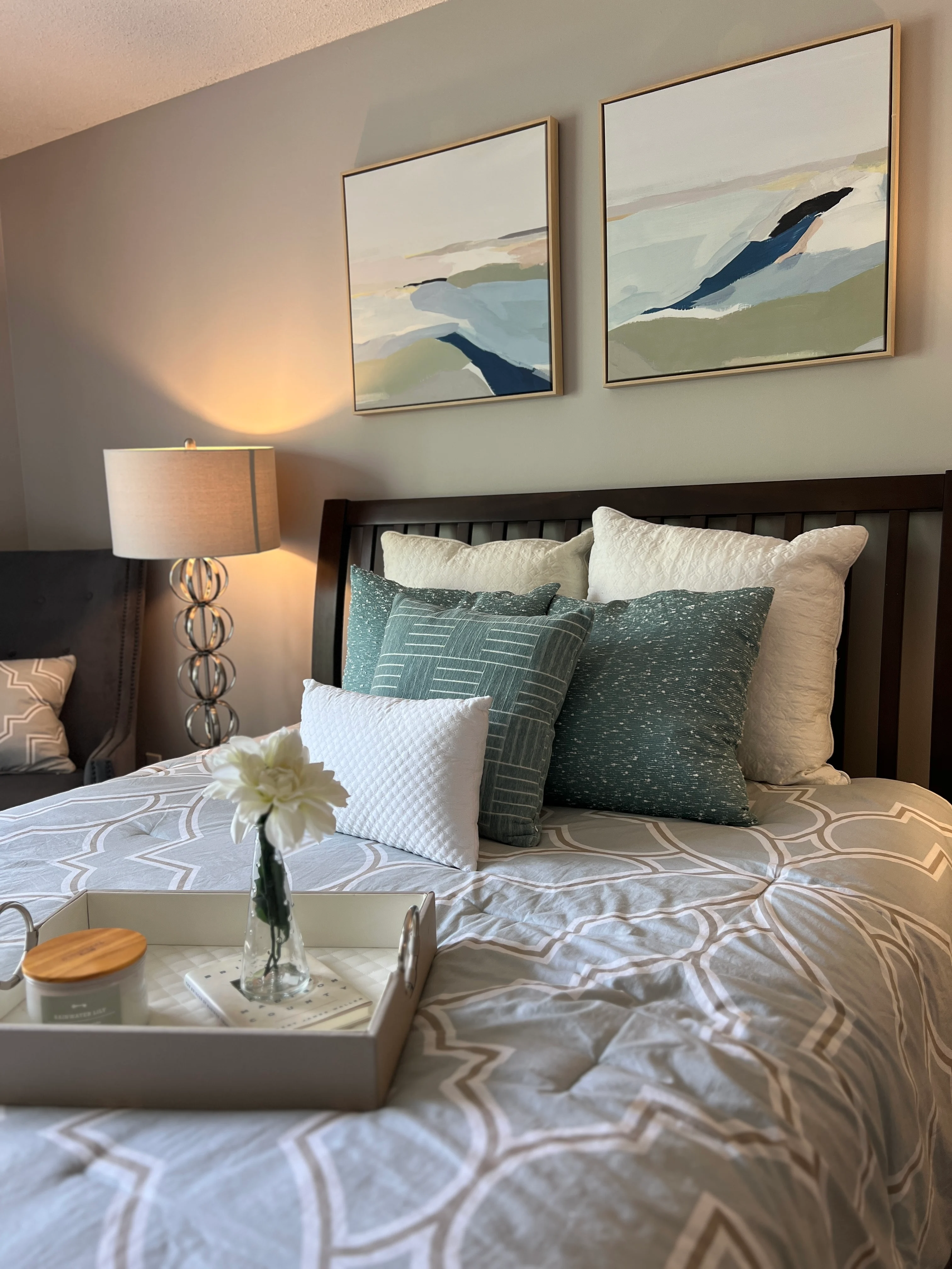 A cozy bedroom featuring a wooden bed with patterned bedding, decorative pillows, and a tray with a flower and snacks on the bed. Two abstract paintings hang on the wall above the bed, illuminated by a nearby lamp.