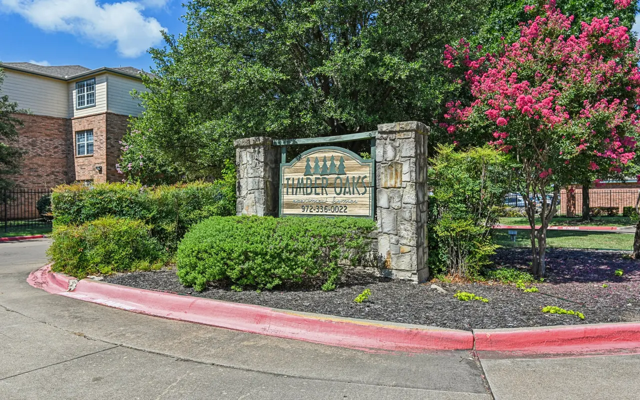 A stone sign for 'Thunder Oaks' surrounded by greenery and blooming trees at an apartment complex entrance. The sign is partially shaded by the trees, and blue skies are visible in the background.
