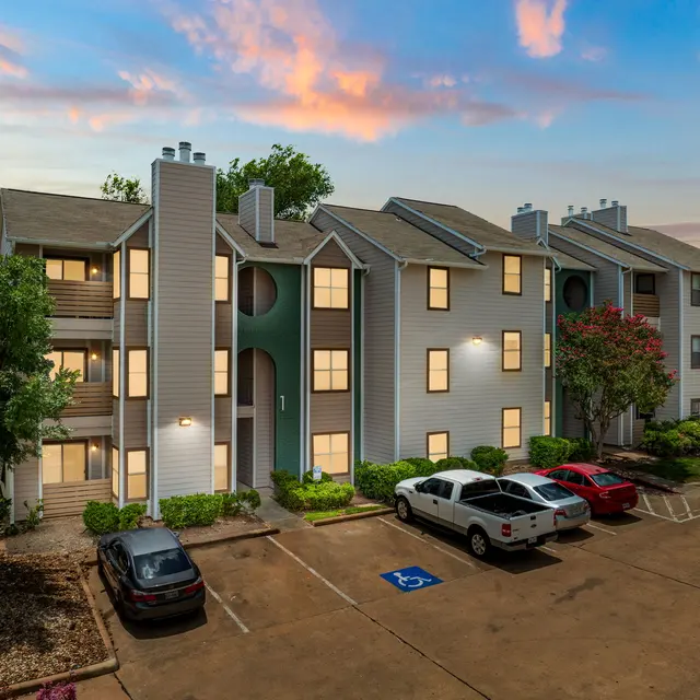 An apartment building during sunset, featuring three stories with balconies and large windows. Cars are parked in front, and trees are visible on the side.