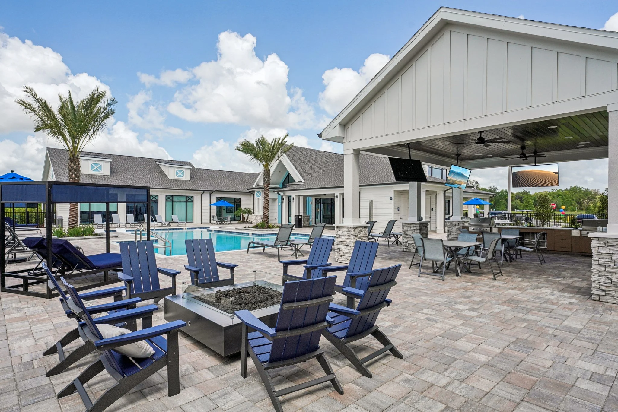 A sunny outdoor pool area featuring blue lounge chairs surrounding a fire pit, with palm trees and a covered seating area nearby.