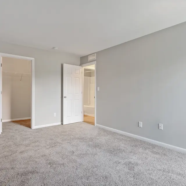 An empty bedroom with light gray walls and carpet. There are two doors visible, one leading to a closet and another outside the room. The space is bright and spacious, with minimal furnishings.