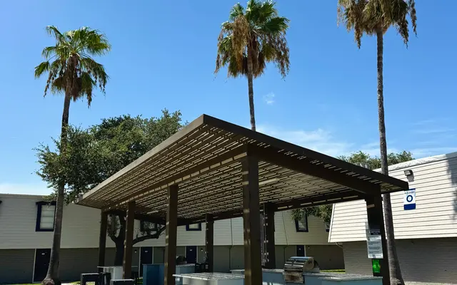 Charming Outdoor Gathering Space An outdoor shelter with a wooden pergola, equipped with bar stools and a countertop, surrounded by lush green grass and palm trees under a clear blue sky.