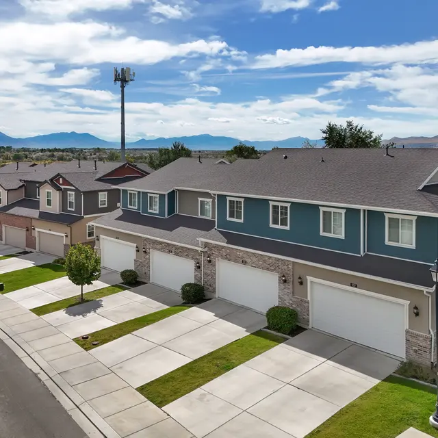 Aerial view of a row of residential homes with neat lawns and driveways, set against a backdrop of mountains and a blue sky with scattered clouds. A cell tower is visible in the background.