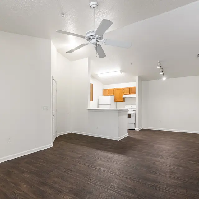 A spacious interior of a modern apartment featuring hardwood flooring, a ceiling fan, and a kitchen area with wood cabinetry.