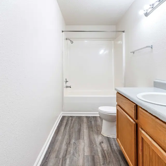 A clean and spacious bathroom featuring a white bathtub and shower combination, a wooden vanity with a sink, and a white toilet. The floor has a modern wood-like appearance and the walls are painted in a light color.