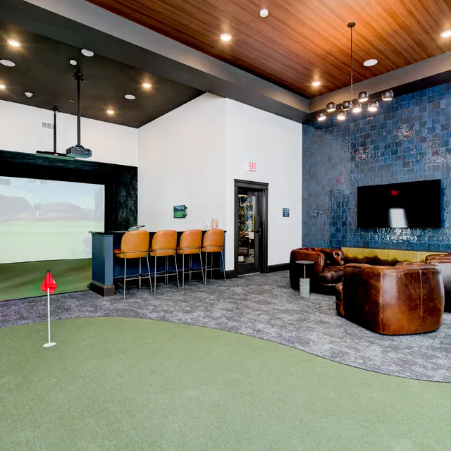 A modern golf simulator room featuring a large screen projector, a bar with high stools, and a seating area with brown leather sofas. The floor is covered with artificial grass and there is a putting green.