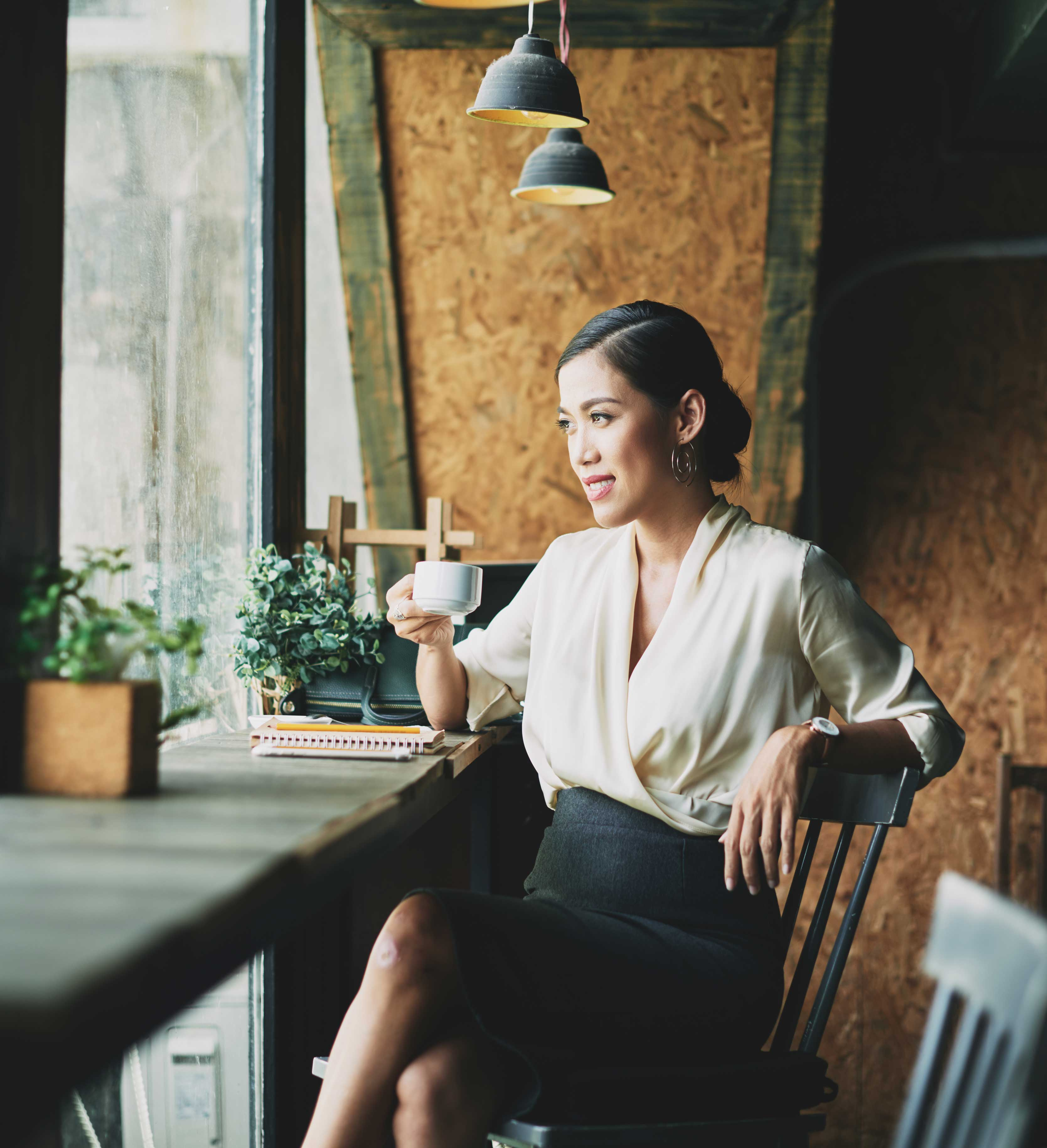 Woman in Cafe A woman sitting in a cafe by a window, holding a coffee cup. She wears a light blouse and a black skirt, with plants and notepads on the table beside her.