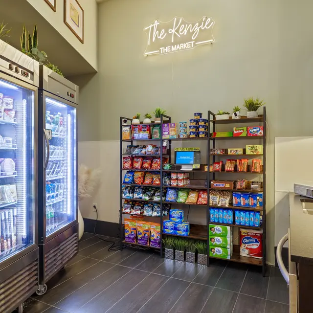 A view of a cozy market area featuring two glass-front refrigerators on the left and a shelving unit with snacks and a digital display on the right. The wall has a sign that reads 'The Kengie The Market'.